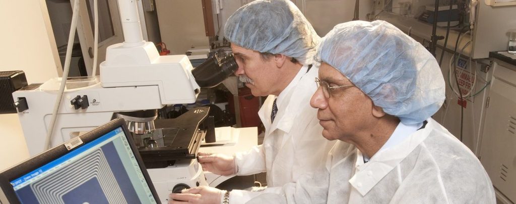 Image of two engineers wearing white lab coats facing left. The engineer on the right is working on a computer, and the engineer on the left is looking through a microscope.