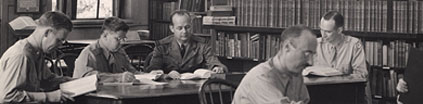 Five men in military uniforms sit at two library tables reading.