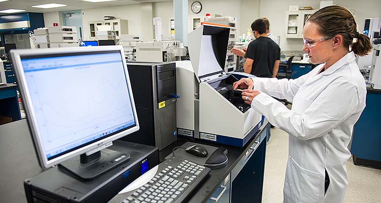 Photo of a woman in a white lab coat and safety glasses placing samples for testing into a small, box-like piece of equipment; and next to the equipment is a computer.