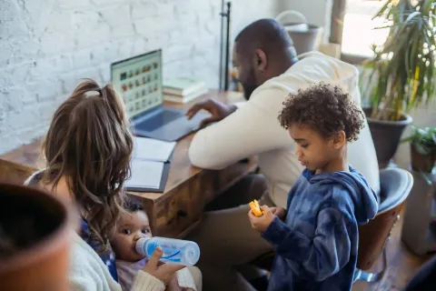 Family at home with father working on laptop, mother feeding baby, and young child playing nearby.