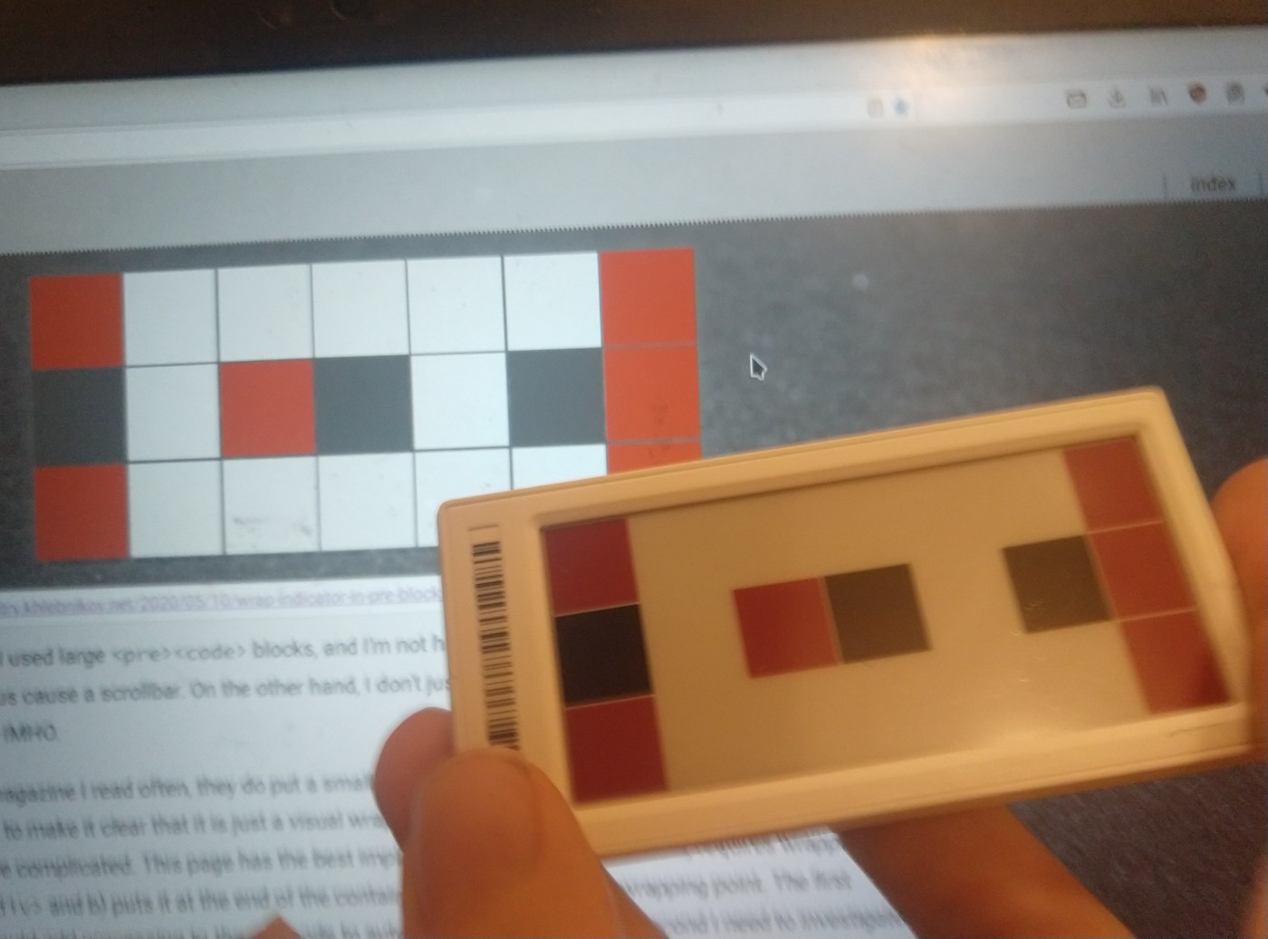 a screen showing the grid at the top of my website. A hand is holding an
e-paper price tag showing the same pattern in front of the screen