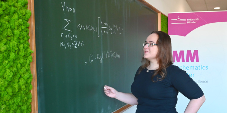 Eine Frau steht vor einer grünen Tafel und schreibt eine mathematische Formel auf.<address