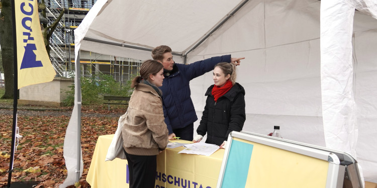 Zu sehen sind eine Studentin und ein Student mit Infomaterial, die einer jungen Besucherin den Weg zu einer Veranstaltung erläutern.<address