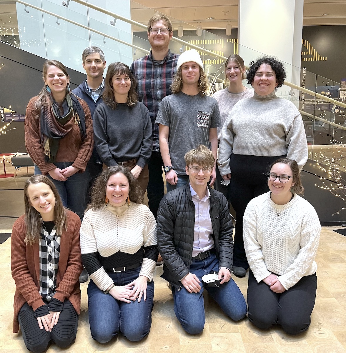 A group of eleven Alaska Sea Grant State Fellows smiles, posing for a group photo in front of a set of stairs inside an office building. Three women and one man is kneeling in the front with three women and one man standing behind them, and two men and one woman in the back. 