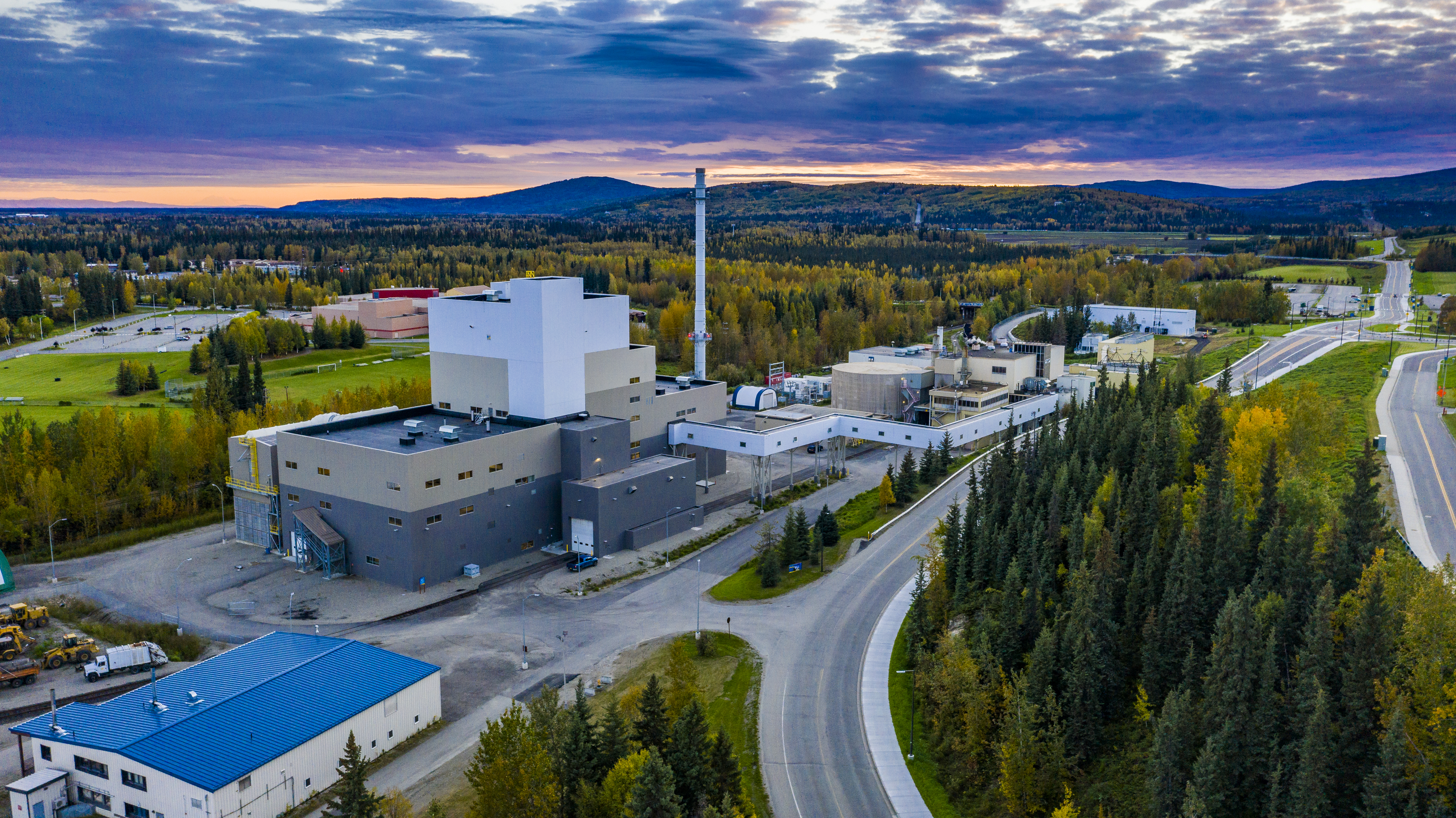 a power plant in the foreground with the sun setting behind hills in the background