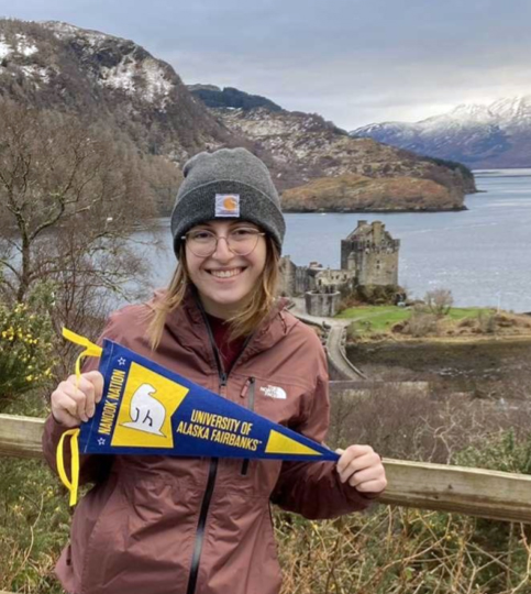 A person with shoulder-length hair and glasses holds a UAF pennant with the seaside in the background.
