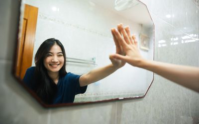 Young adult smile asian woman practice self talk conversation in the morning in bathroom at home. Hand hi-touch at mirror. Healthy lifestyle after wake up concept.