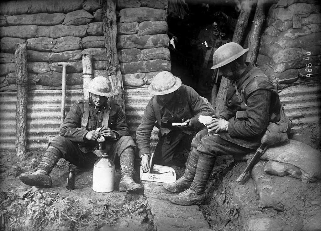 A photograph of three allied soldiers sitting together for a meal in a trench wearing camouflage uniforms and metal helmets.