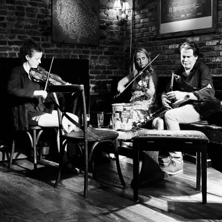 Two women playing fiddle and a man playing pipes sitting around a table in the corner of a bar with exposed brick walls.