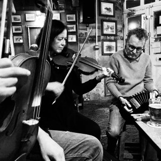 A woman playing fiddle and a man playing concertina in an ornate pub. In the foreground another man holds a fiddle.