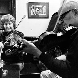 Two fiddlers, a man and a woman, in the corner of a pub.