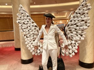 Sue in a white suit in front of angel wings made of pointe shoes.