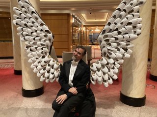 Dan in tux in front of angel wings made of pointe shoes.