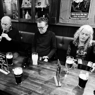 A man playing whistle, a man playing concertina and a woman playing concertina all lined up sitting with their backs to the wall in front of a pub table.