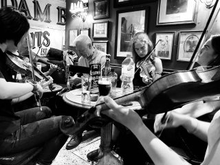 Loads of fiddlers gathered ’round a piper playing in a pub.