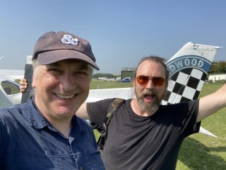 A selfie of me smiling with Andy goofing around in front of our light aircraft on a green airfield.