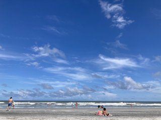 Waves breaking on a sandy beach where some people are standing or lying under a blue sky dotted with a few clouds.