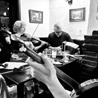 In the foreground a hand grips a banjo neck while in the background a woman plays fiddle and a man plays accordion.