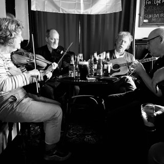 Two fiddlers and a tin whistle player accompanying a man singing and playing guitar, all of around a small pub table festooned with drinks.