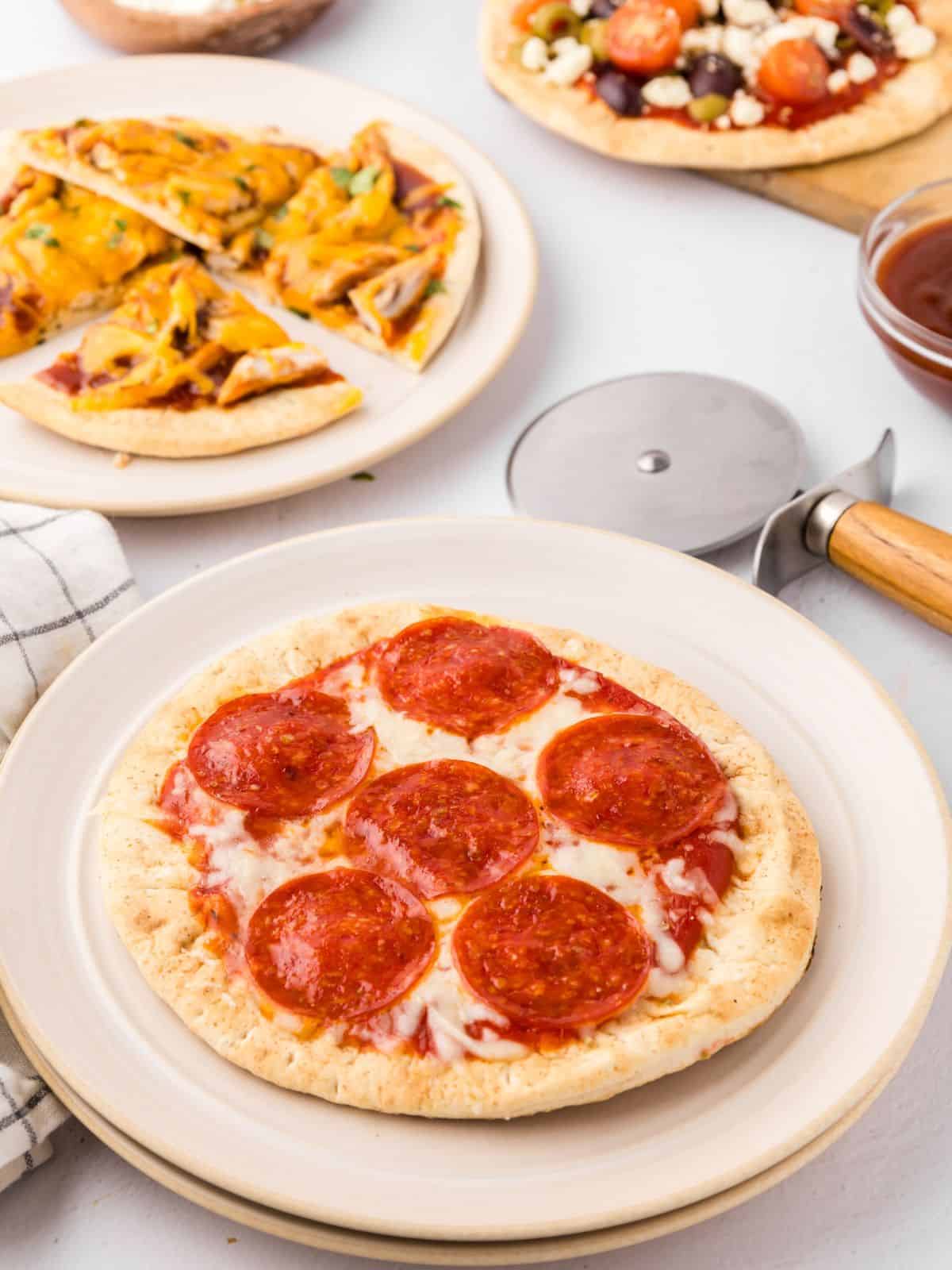 A variety of assembled and baked pita pizzas on white plates next to pizza cutter and fresh parsley.