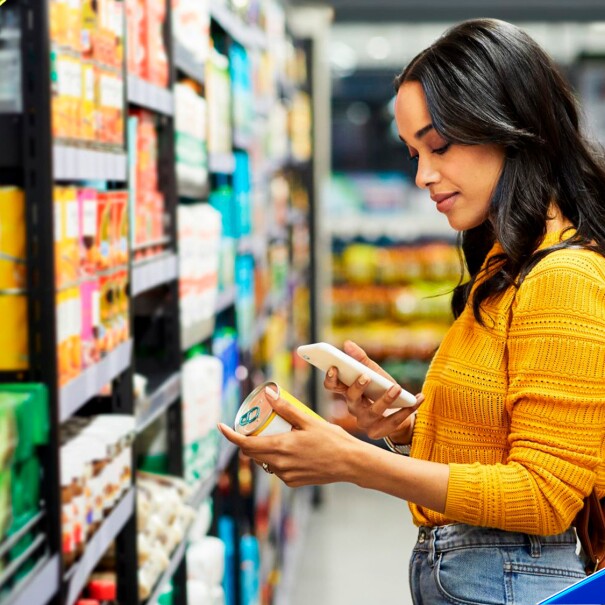 A woman stands in a grocery store aisle and holds a can of food while looking at her phone.