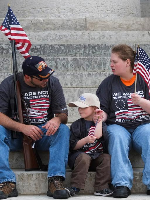 Eine junge Familie mit amerikanischer Flaggen sitzt auf Stufen vor einem Gebäude. Der Vater lehnt an einem Gewehr, sie unterhalten sich.