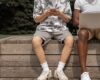 Two men using mobile devices and laptop sitting on a bench