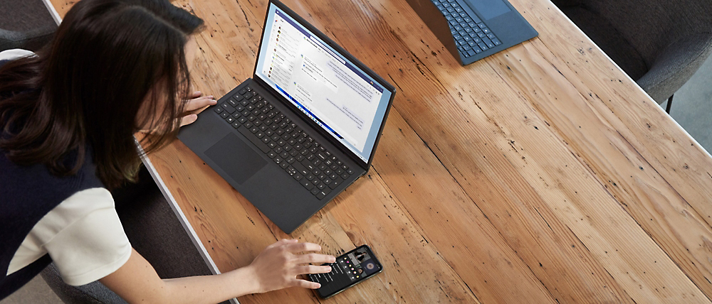 A woman works on a laptop at a wooden table, also using a handheld device, viewed from above.