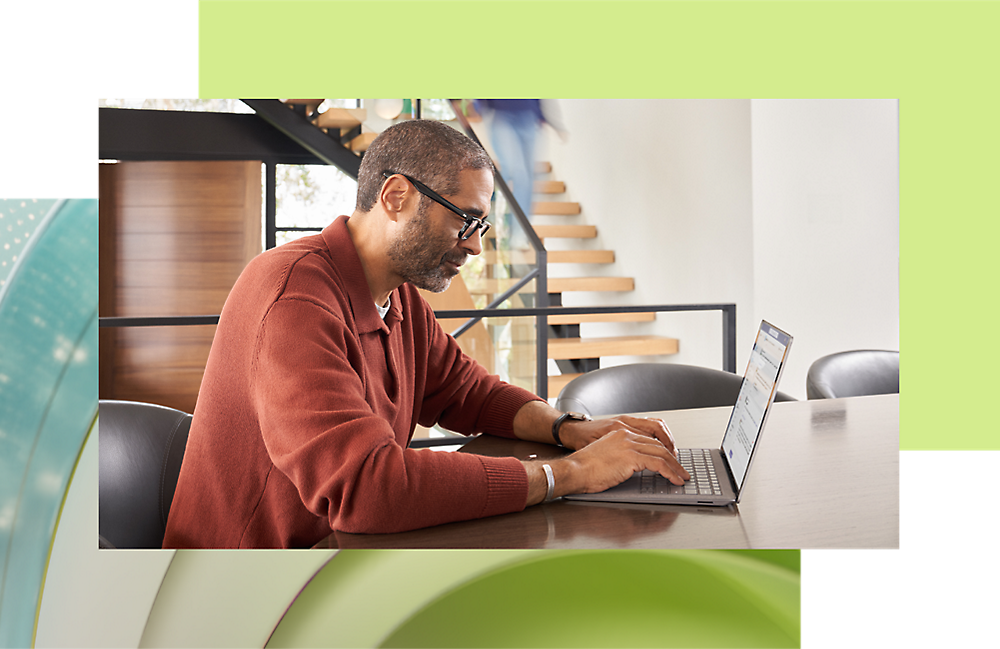 A man wearing glasses and a red cardigan works on a laptop at a modern desk in a brightly lit office space.