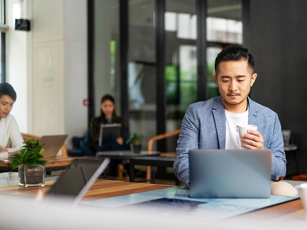 Seorang pria dengan blazer biru duduk di depan meja dengan laptop dan memegang cangkir putih. 