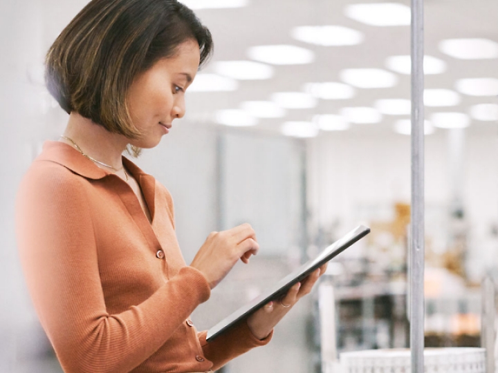 Eine Frau in einem orangefarbenen Oberteil verwendet ein Tablet, während sie in einem gut beleuchteten Raum steht, möglicherweise in einem Büro oder einem Labor.