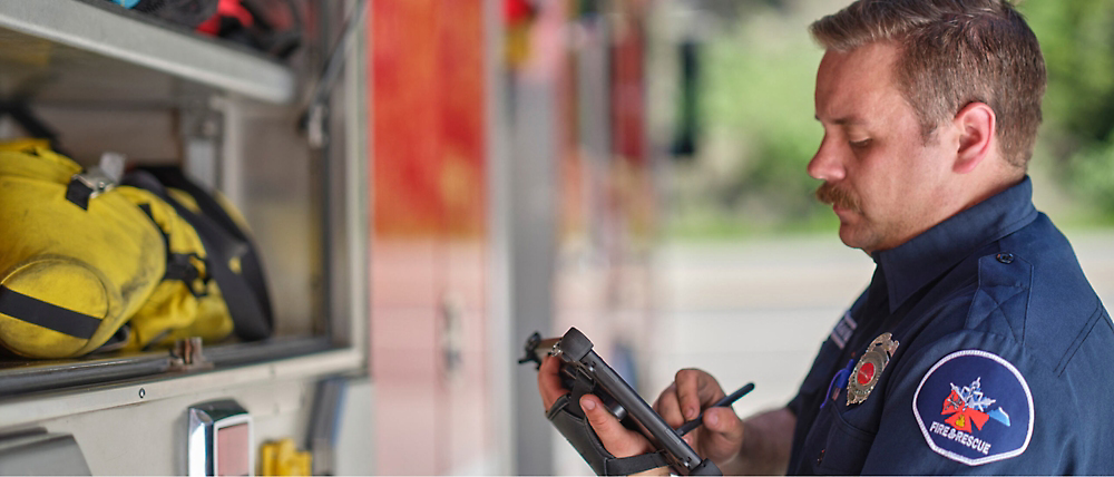 A firefighter in uniform reviews information on a tablet beside a fire truck equipped with emergency gear.