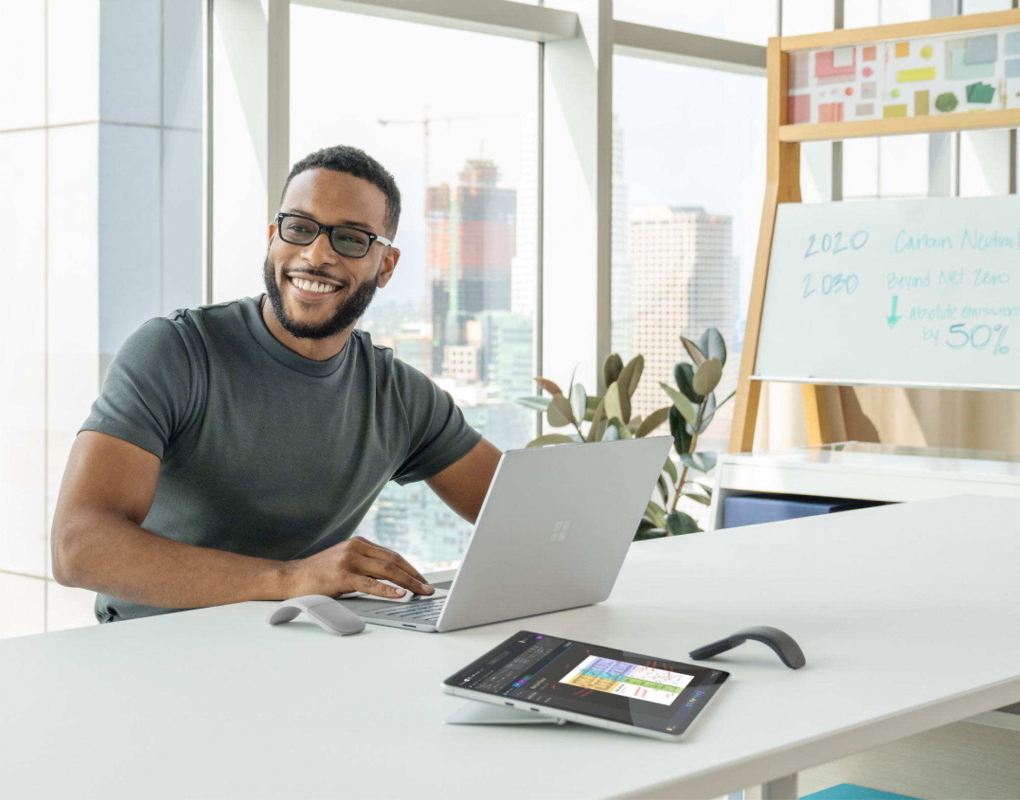 Smiling man in glasses using laptop at a modern office desk with a tablet, phone, and presentation board