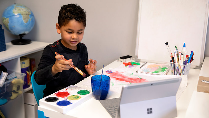  A young student, painting at home, using a Surface Go for remote learning.