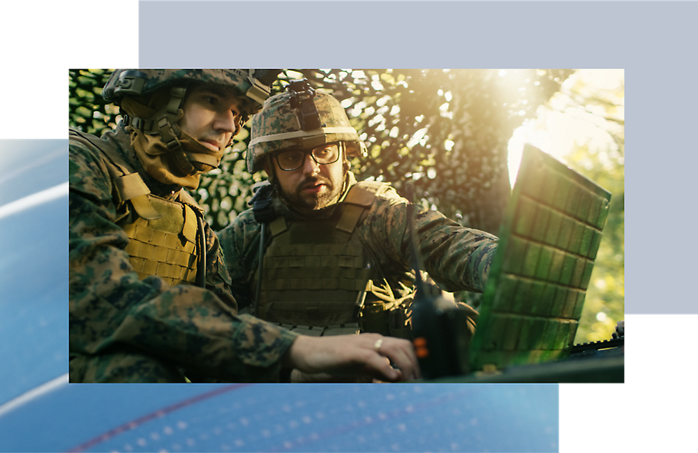 Two soldiers in camouflage examining a computer in a forest setting with sunlight filtering through leaves.