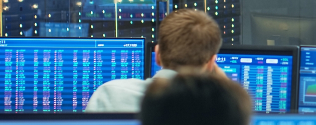 Back view of two people monitoring financial data on multiple blue screens in a control room environment.