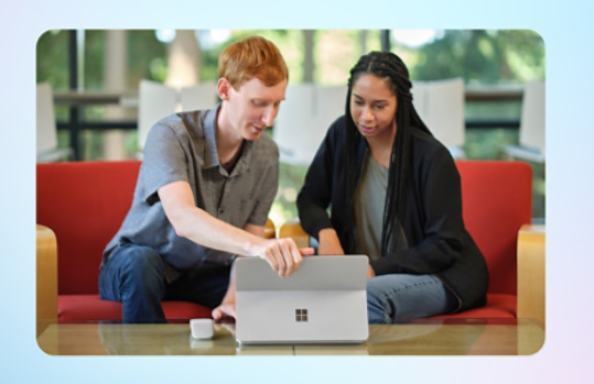 Two students are looking at the screen of a platinum Surface computer. A light grey Surface Arc Mouse sits on the table.