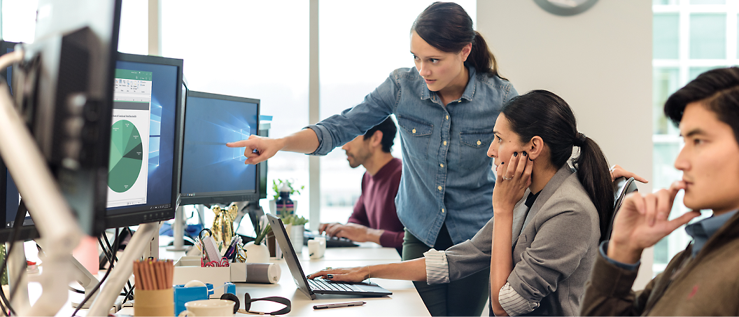 A woman points at a computer screen while another woman and two men work at their desks in an office.