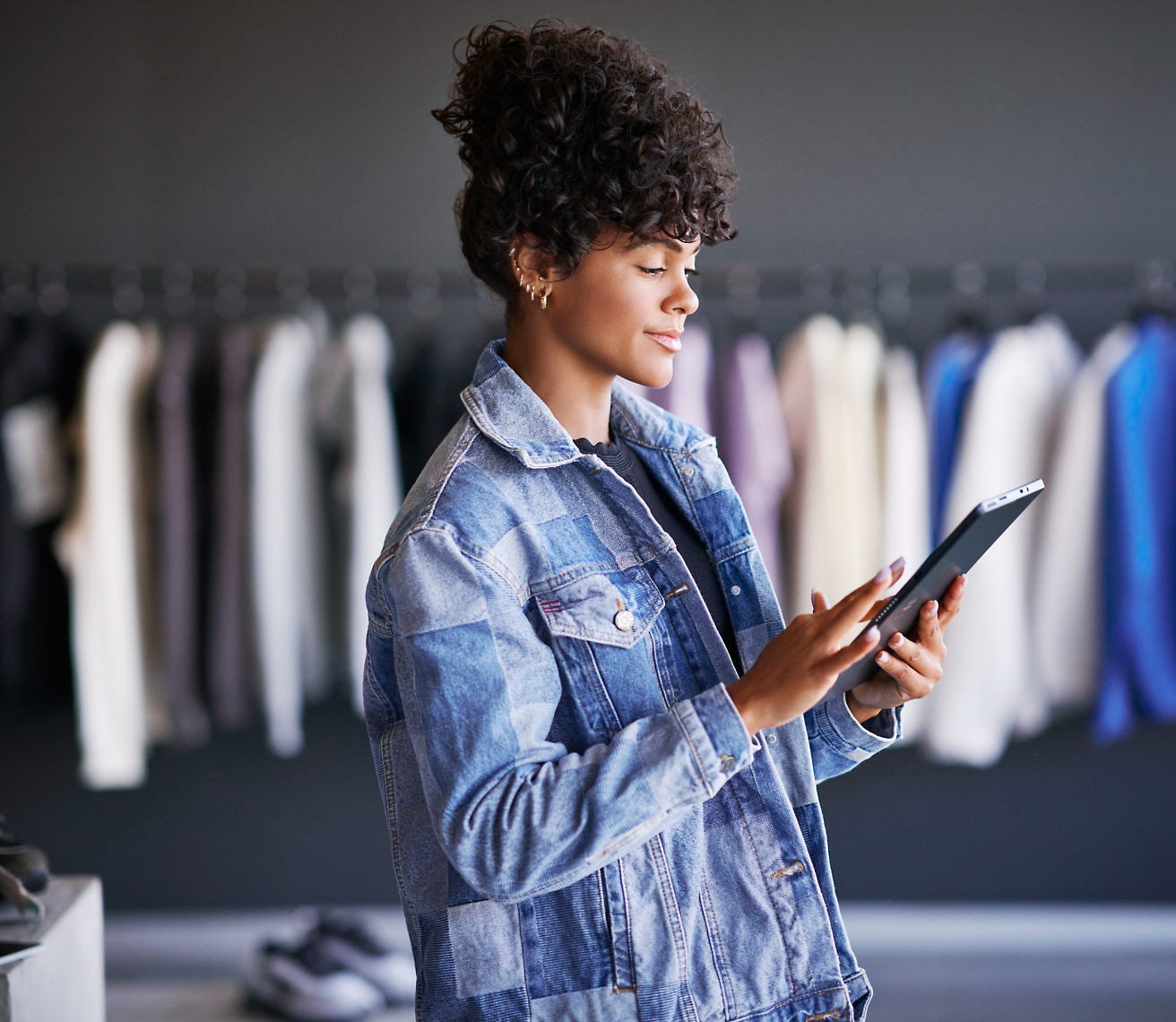 A person wearing a denim jacket is using a tablet in a clothing store with various garments hanging on racks in the background.