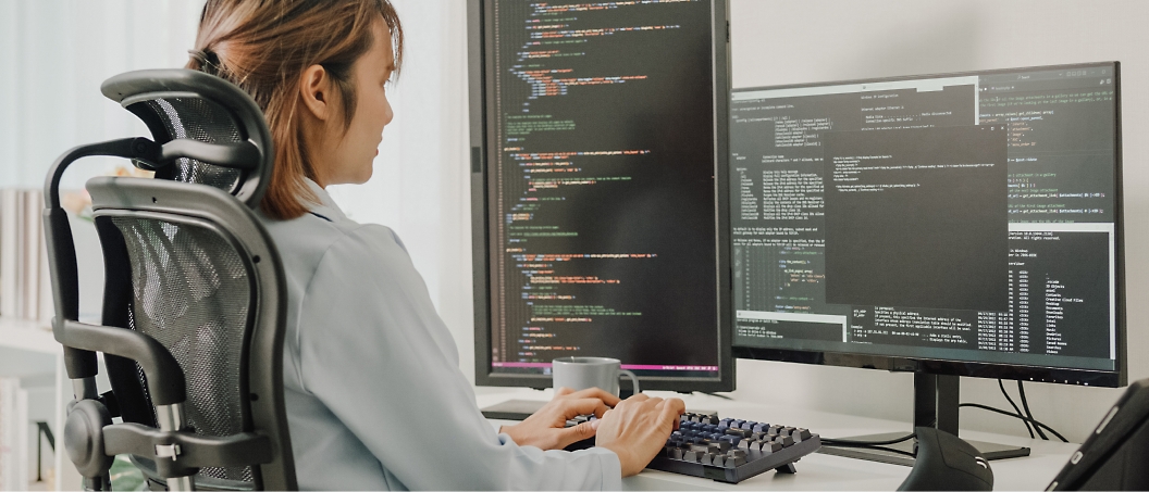 A person sits at a dual-monitor setup in an office chair, typing code on their keyboard