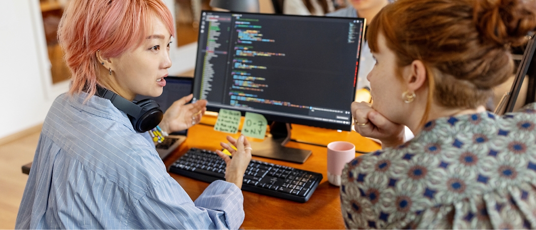 Two people discussing code displayed on a computer monitor at a workstation with a keyboard, headphones, and a mug.