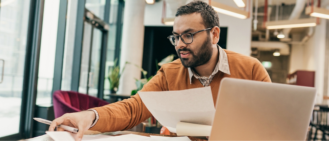 Man in an office setting, wearing glasses and an orange sweater, looks at papers in one hand while using a laptop.