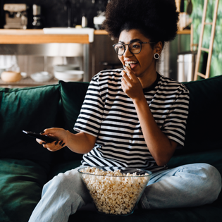 A woman sits on the couch, eating popcorn and watching TV.