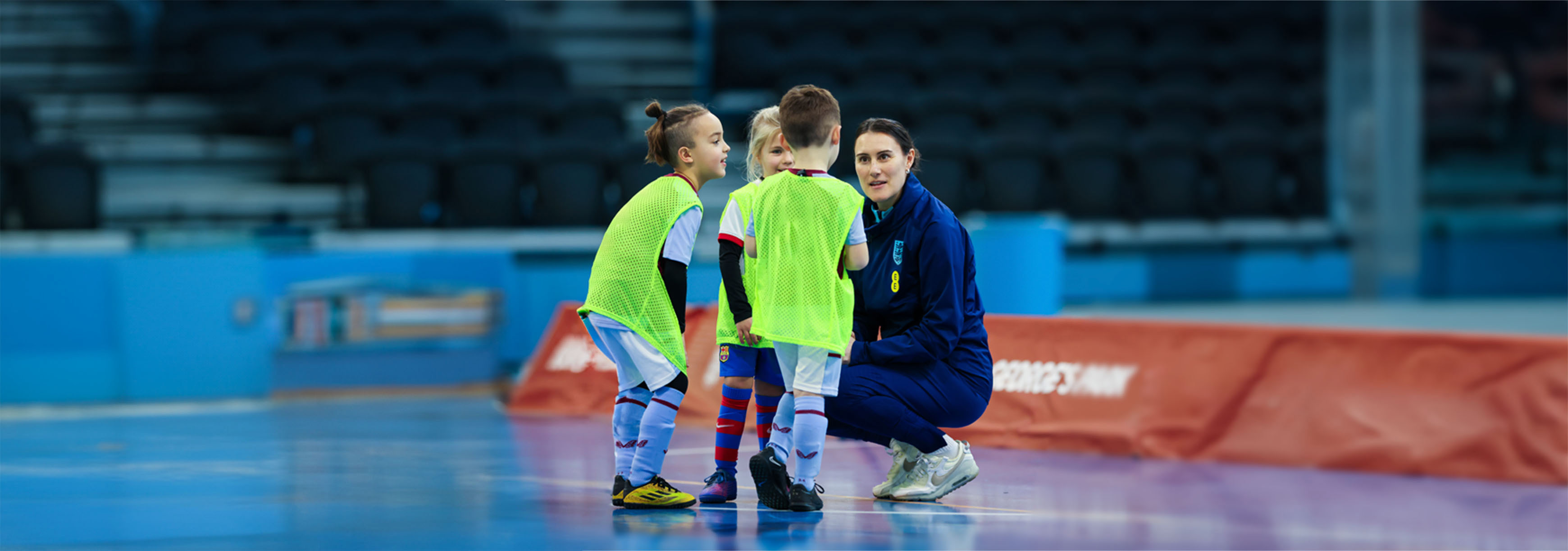 During a training session on a futsal court, an FA coach smiles and listens to three young players.