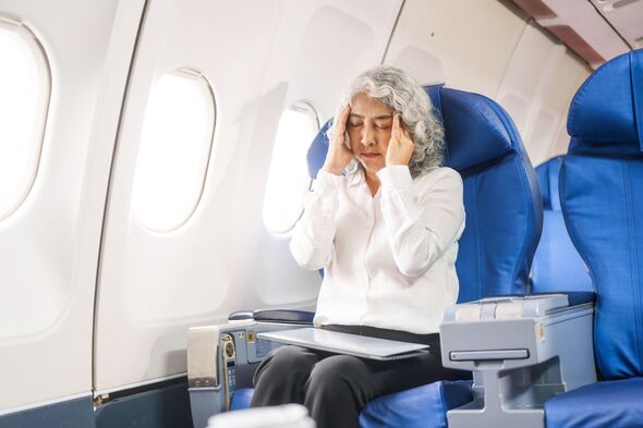 A mature woman sits by the window on a passenger plane, traveling abroad. She looks unwell, experiencing a headache, nausea, and vomiting, which dampe