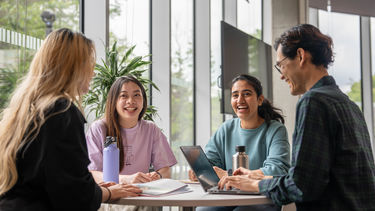 Four smiling postgraduate students sat working together in a campus location - one of the group is typing on their laptop. 