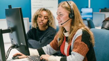 Students on computers, one of which has a headset and is talking to a client