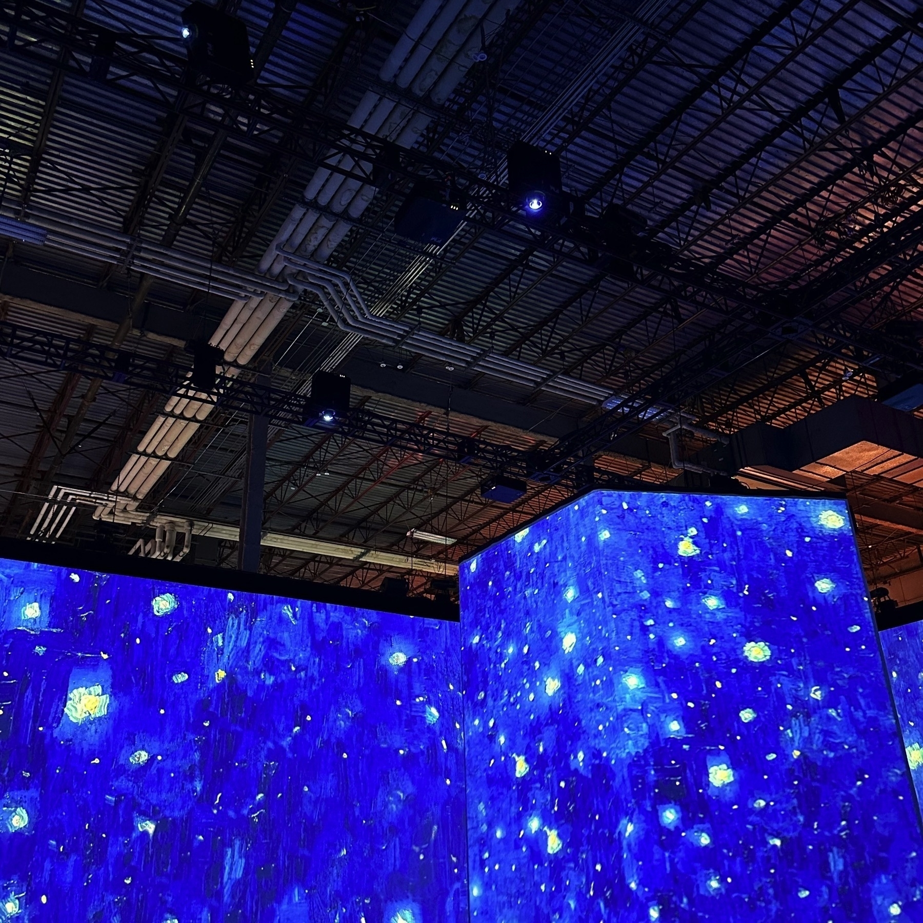 A large room is illuminated with dynamic, starry blue projections reminiscent of Van Gogh's Starry Night, with pipes visible on the ceiling.