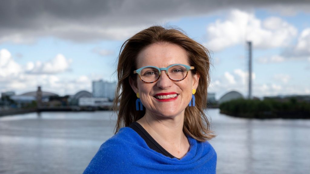 Headshot of Catherine Stihler, wearing a blue shawl standing in front of a body of water with buildings and clouds in the distance.