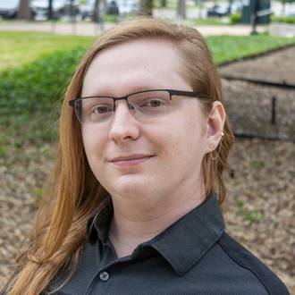 Tyler is facing forward and smiling, He has red hair and wears glasses and a black shirt.